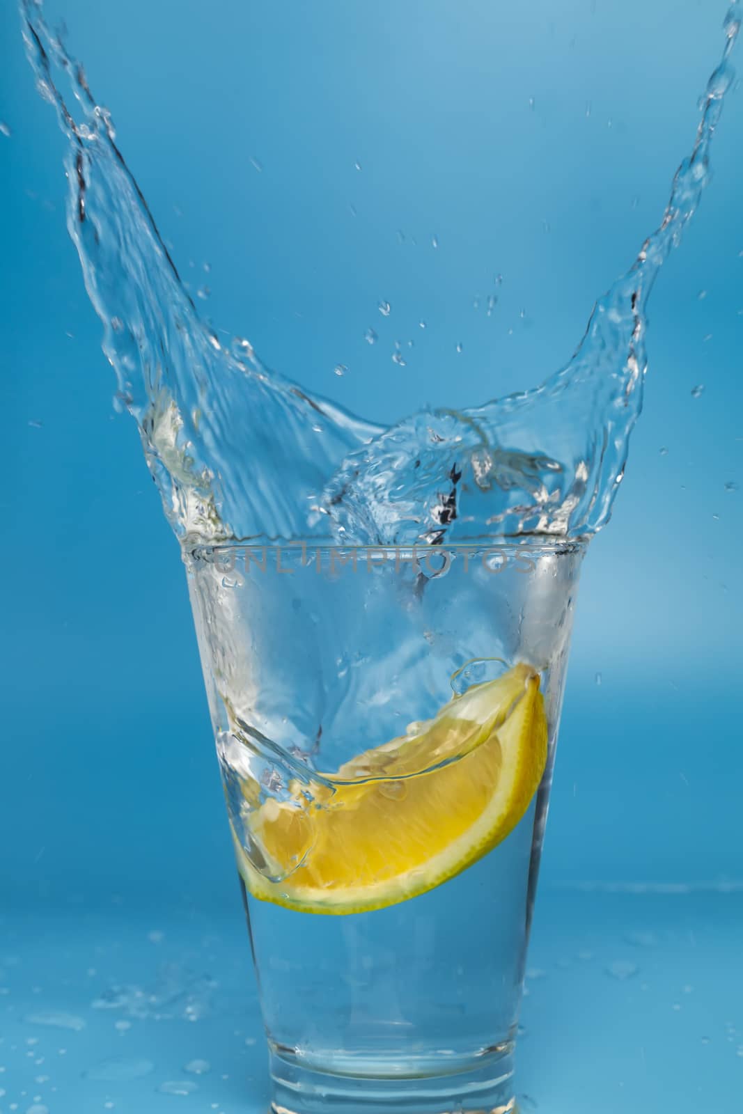Lemon slice falling and dropping into a splashing glass with light blue background.