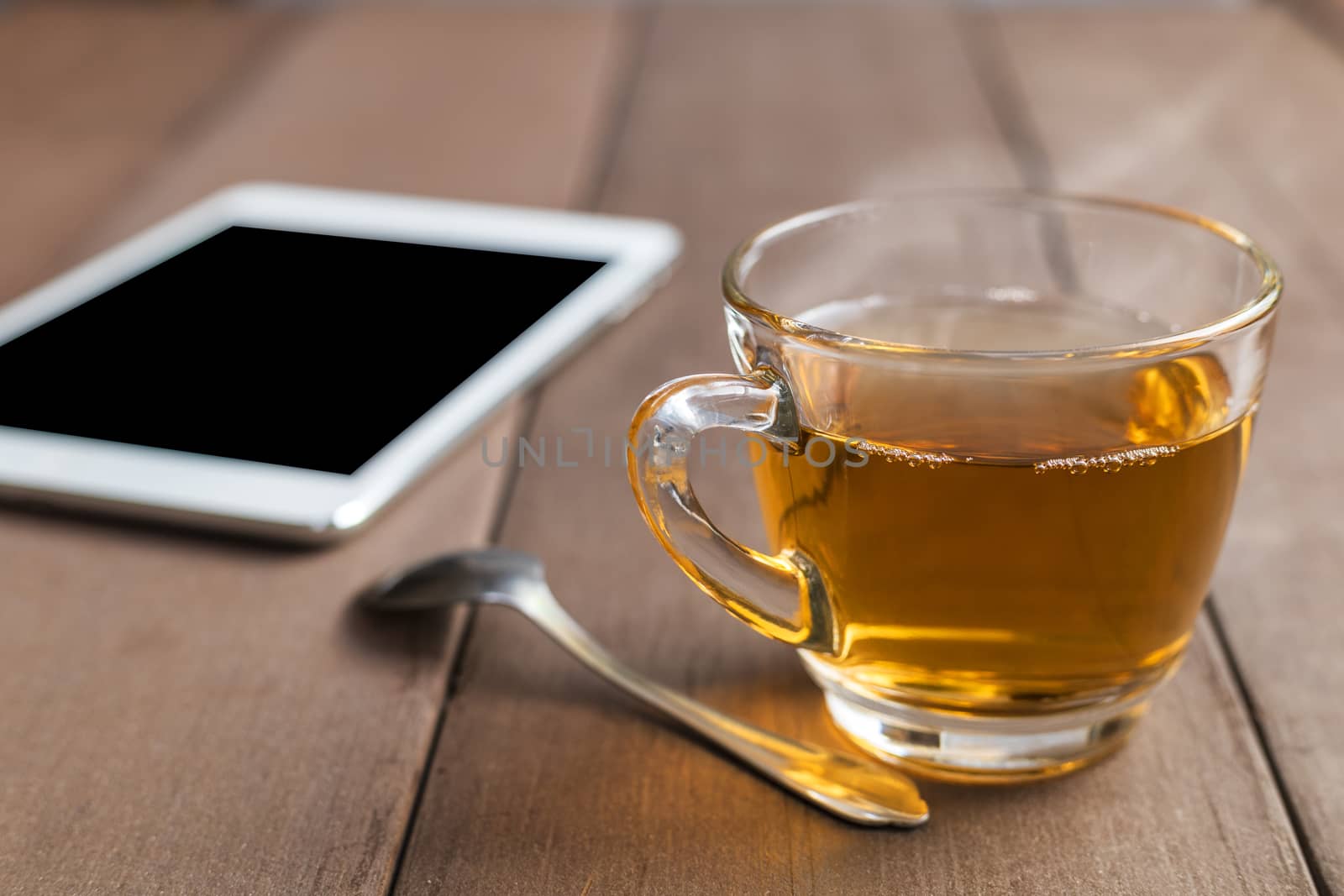 Close up hot tea cup on wood table with tea spoon and tablet shot on morning time with sun ray background. Soft focus and selective focus to tea cup. hot tea in glass with copy space for text or design.