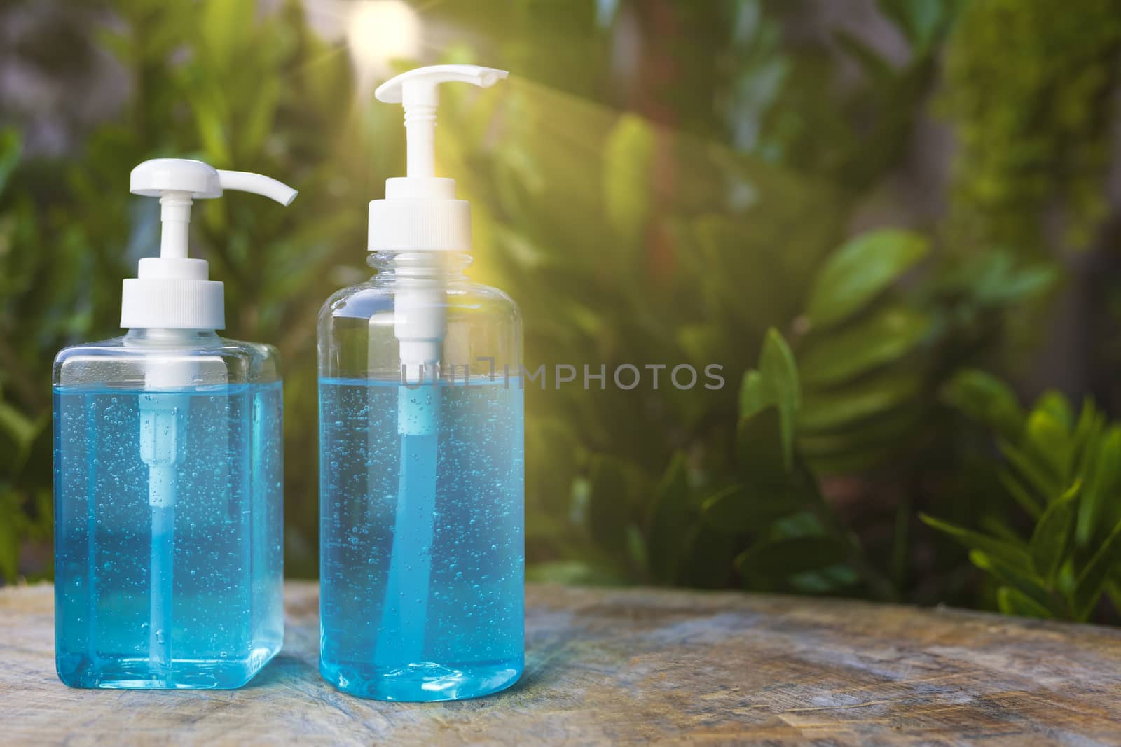 Blue alcohols gel bottle on blur green nature background and sun ray. Hand sanitizer gel in pump bottle. With copy space for text or design. Blue alcohols gel on wood table. Alcohols gel in 2 different size pump bottle. 