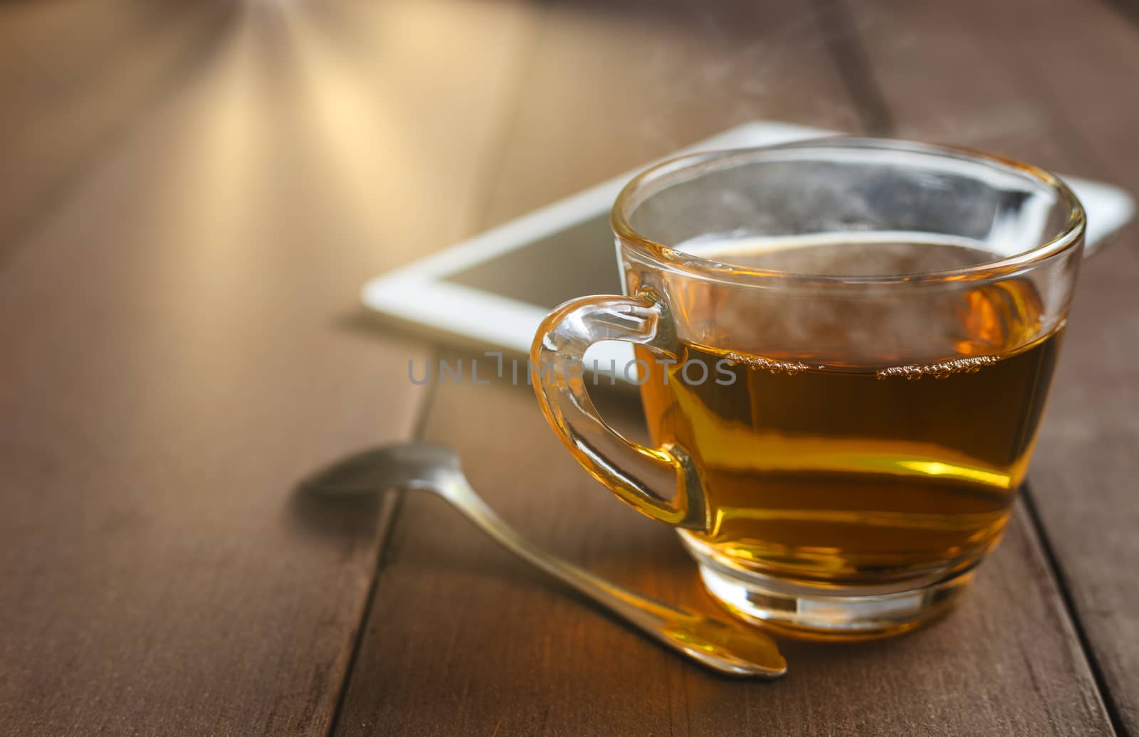 Close up hot tea cup on wood table with tea spoon and tablet shot on moning time witn sun ray background. Soft focus and selective focus to tea cup. hot tea in glass with copy space for text or design.