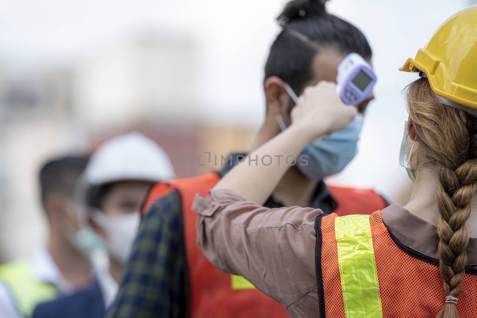 Factory woman worker in a face medical mask and safety dress used measures temperature at worker people standing on queue with a non-contact infrared thermometer.
