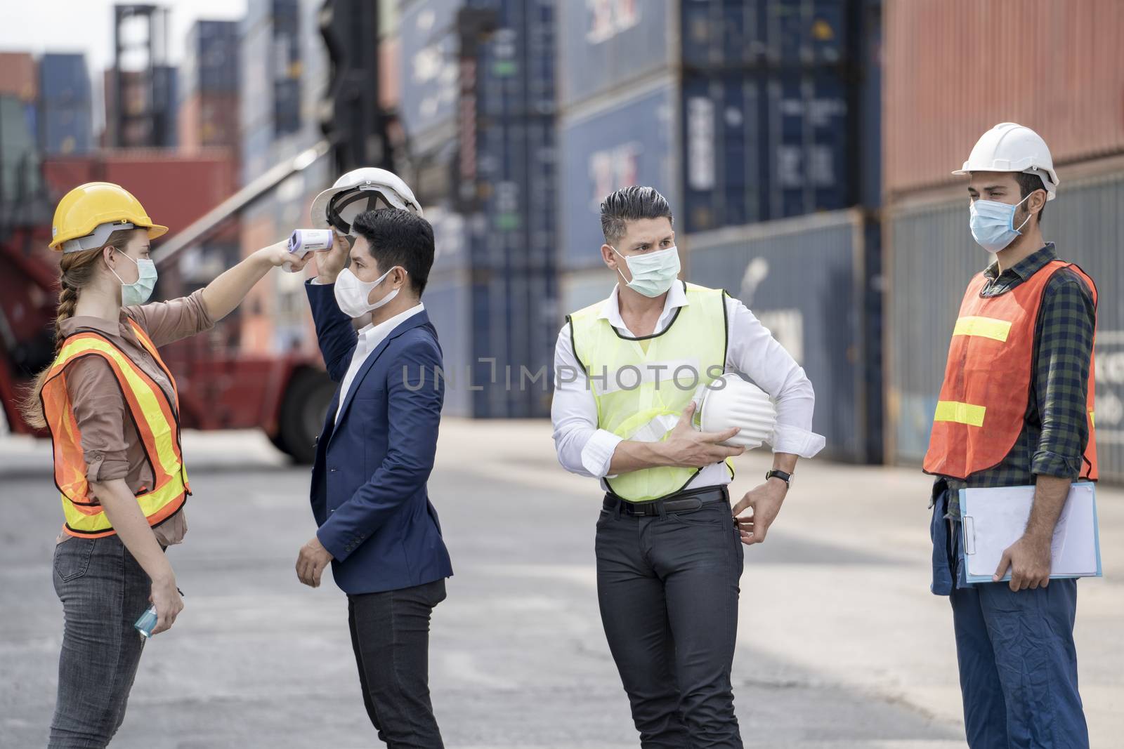 Factory woman worker in a face medical mask and safety dress used measures temperature at worker people standing on queue with a non-contact infrared thermometer.

