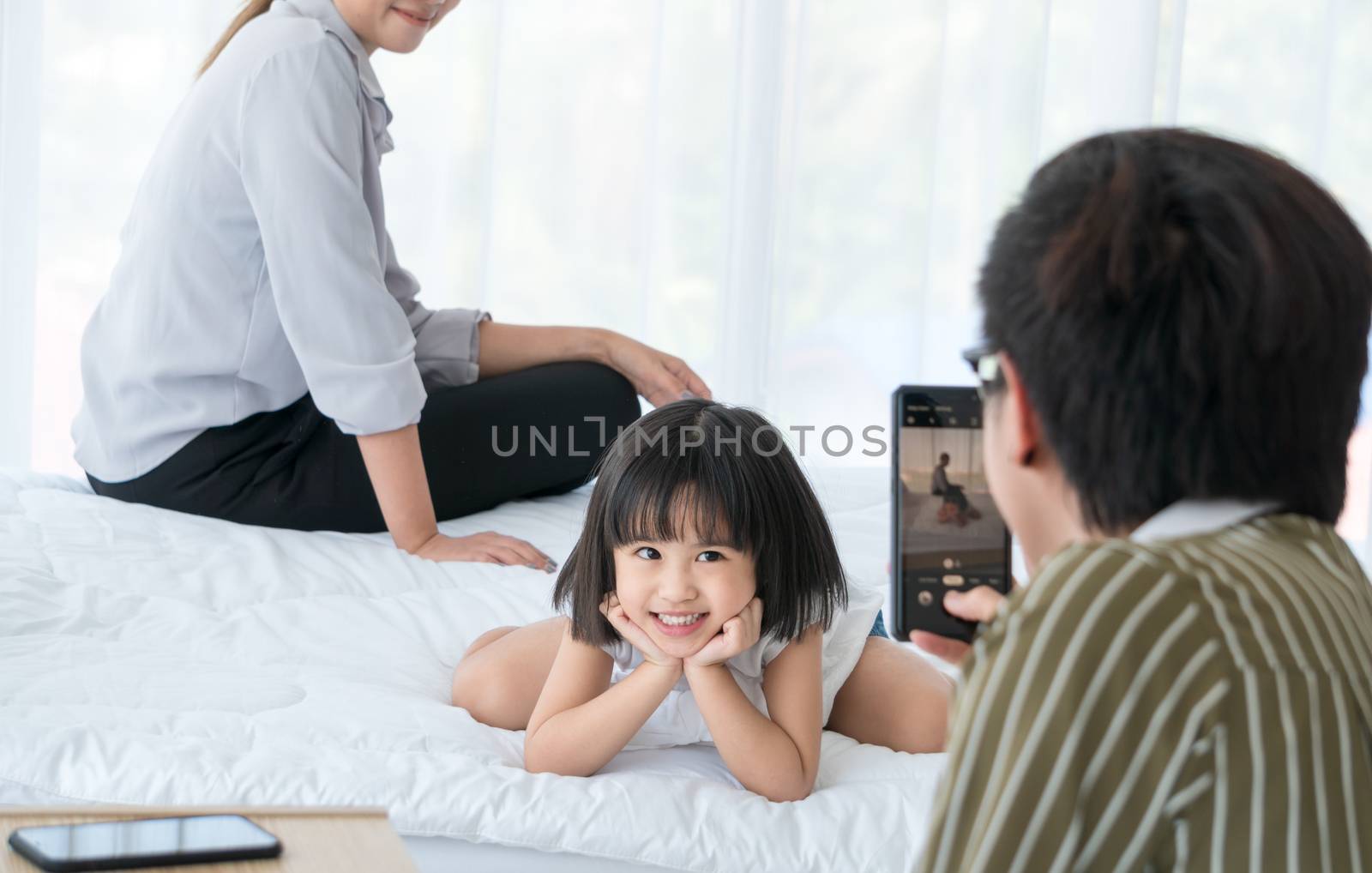 Asian family with cute parents and daughter. The father was using a cell phone to take pictures of his daughter and mother sitting nearby. They are having fun playing in the bedroom.