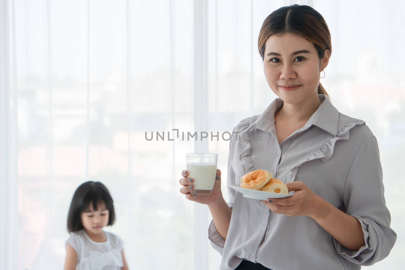 Asian mum is happily living with her little daughter. She holds a glass of milk and a bread plate for her daughter.