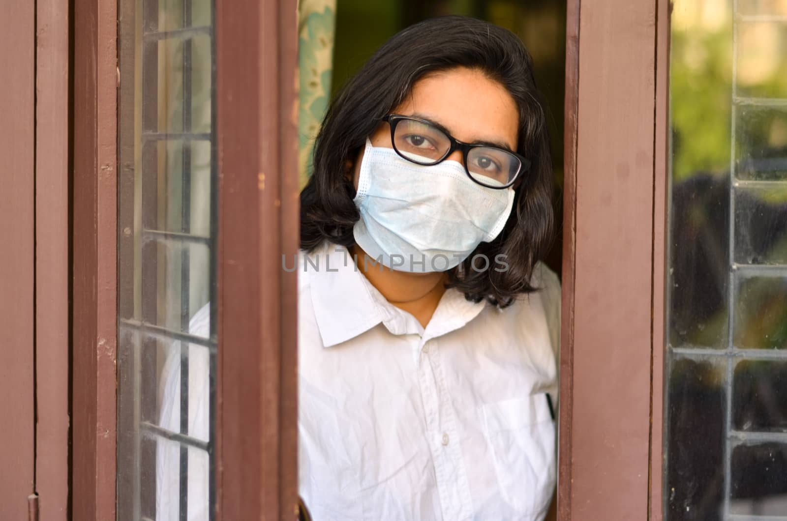 Concerned & worried lady under lock down home quarantine wearing protective breathing surgical face mask against flu and novel Corona virus (Covid-19) pandemic & greeting visitor through door / window