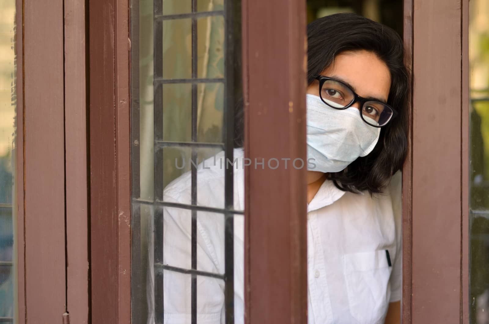 Concerned & worried lady under lock down home quarantine wearing protective breathing surgical face mask against flu and novel Corona virus (Covid-19) pandemic & greeting visitor through door / window