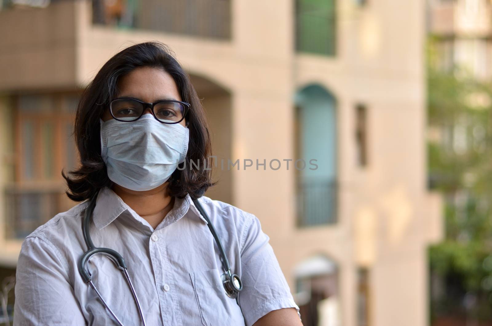 Portrait of young medical healthcare female worker with hands crossed / folded, stethoscope over her shoulders wearing surgical mask to protect herself from Corona Virus (COVID-19) pandemic disease