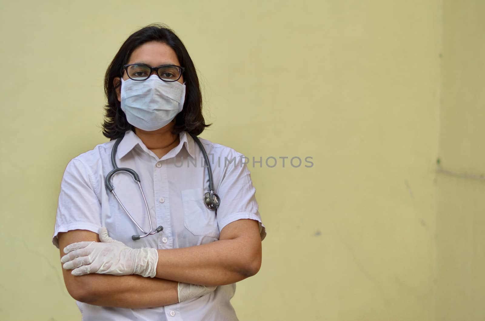 Portrait of a young medical healthcare female worker with hands crossed / folded, wearing surgical mask to protect herself from Corona Virus (COVID-19) pandemic disease against yellow background