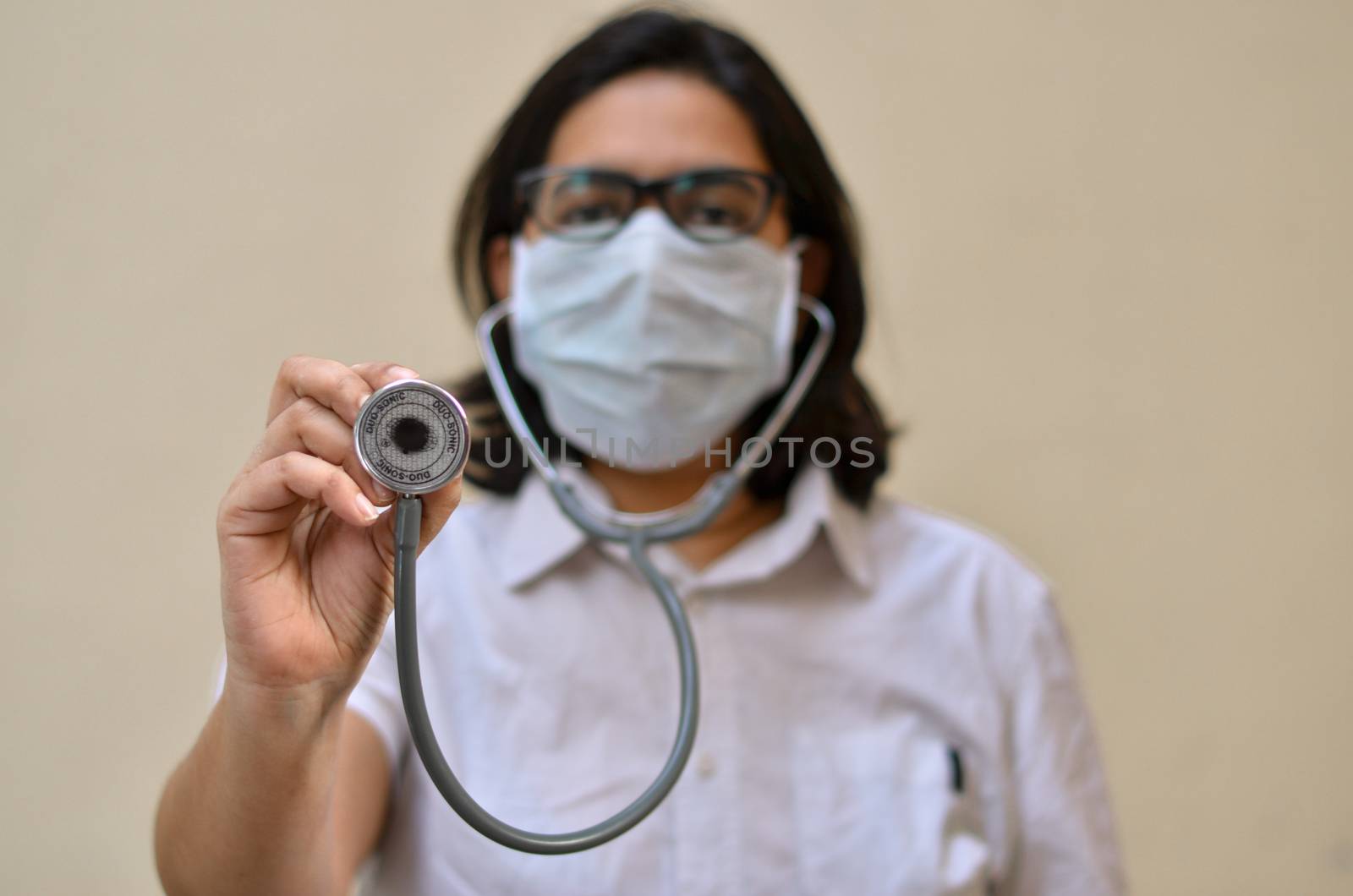 Portrait of young medical healthcare female worker showing / holding out her stethoscope & wearing surgical face mask to protect from Corona Virus (COVID-19) pandemic against yellow background