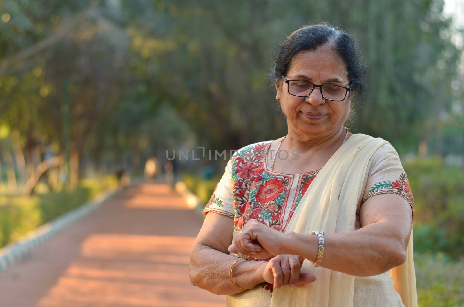 Senior Indian woman smiling and looking at her hands / smart watch to know the time in a park. She's wearing an off white salwar kamiz. Concept - Digital literacy in India for senior citizens