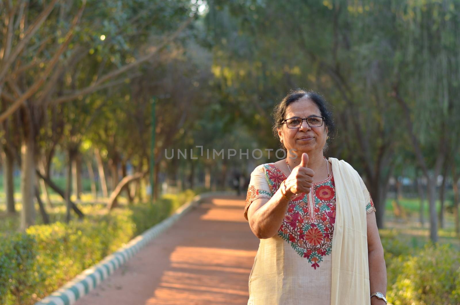 Portrait shot of a happy looking senior north Indian woman wearing traditional chikan kari Indian salwar kameez showing a thumbs up in a garden against a bokeh of canopy of trees.