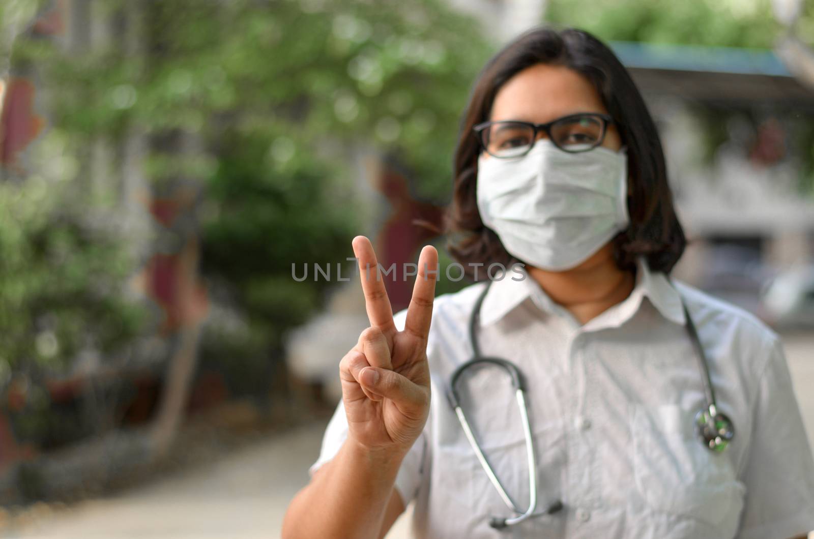 Portrait of a young medical healthcare female worker making a victory sign with her right hand wearing surgical mask to protect herself from Corona Virus (COVID-19) pandemic stethoscope around neck