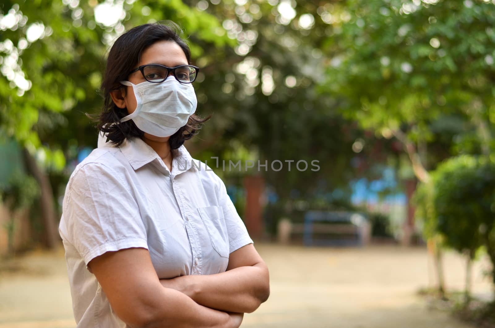 Side angle portrait of a young medical healthcare female worker with hands crossed / folded, wearing surgical mask to protect herself from Corona Virus (COVID-19) pandemic against blur background