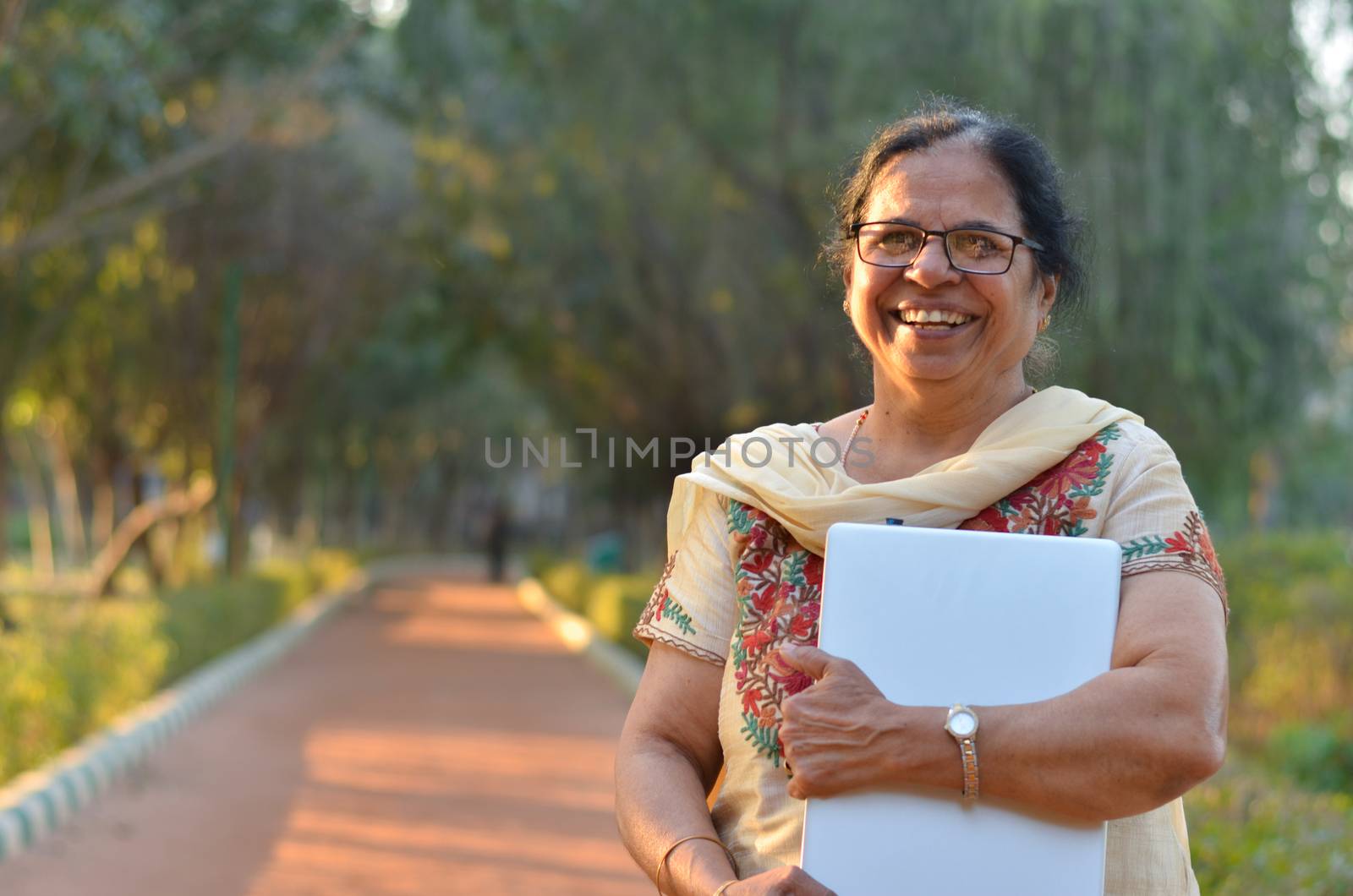 Portrait of a confident retired Senior Indian woman carrying a laptop in a park wearing off white salwar kamiz. Concept - Digital literacy in India for senior citizens