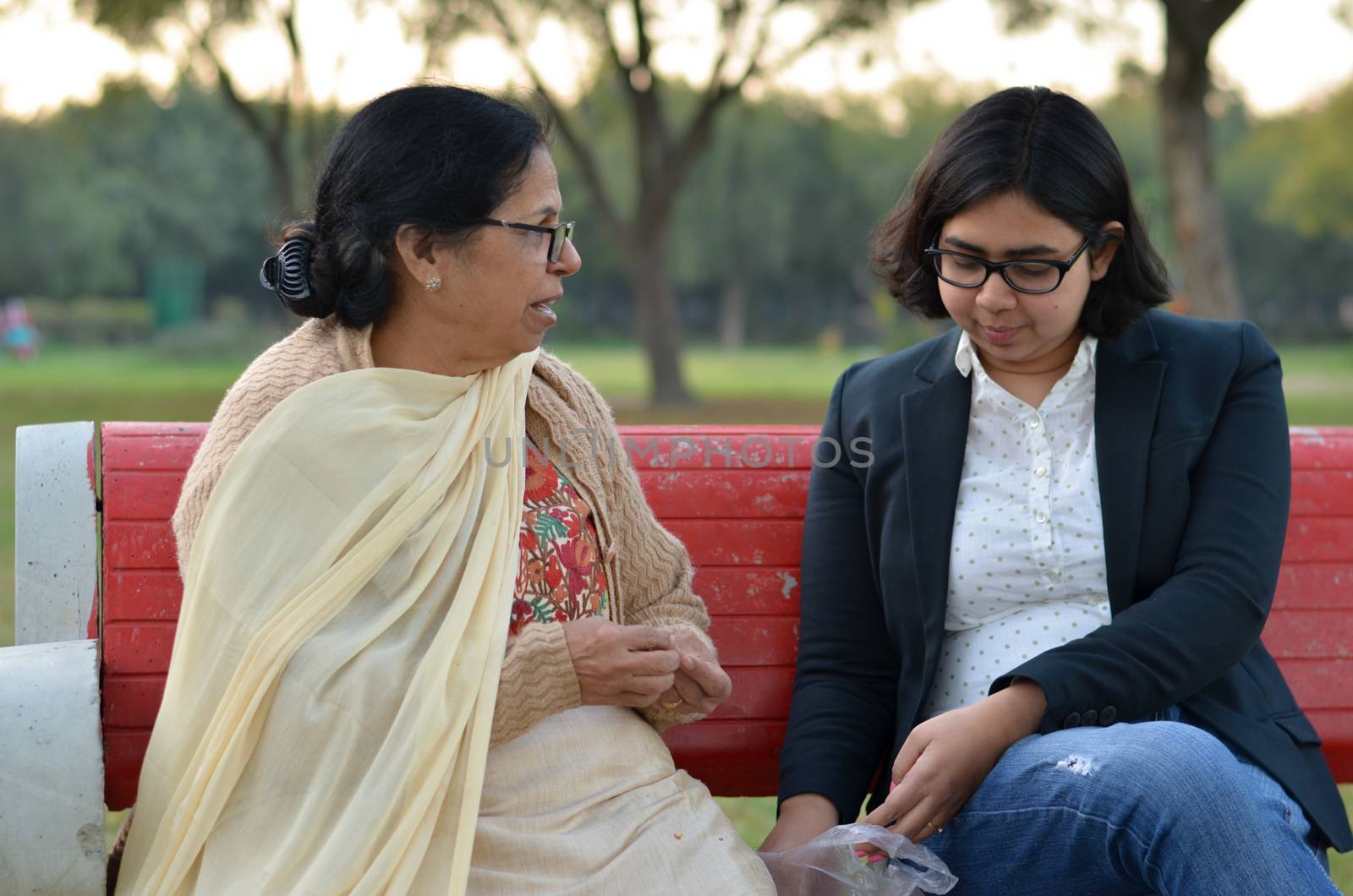 Shot of a senior retired Indian woman sitting in a park with her daughter in law on a red bench peeling peanuts, and eating them. Concept - Happy Retired life