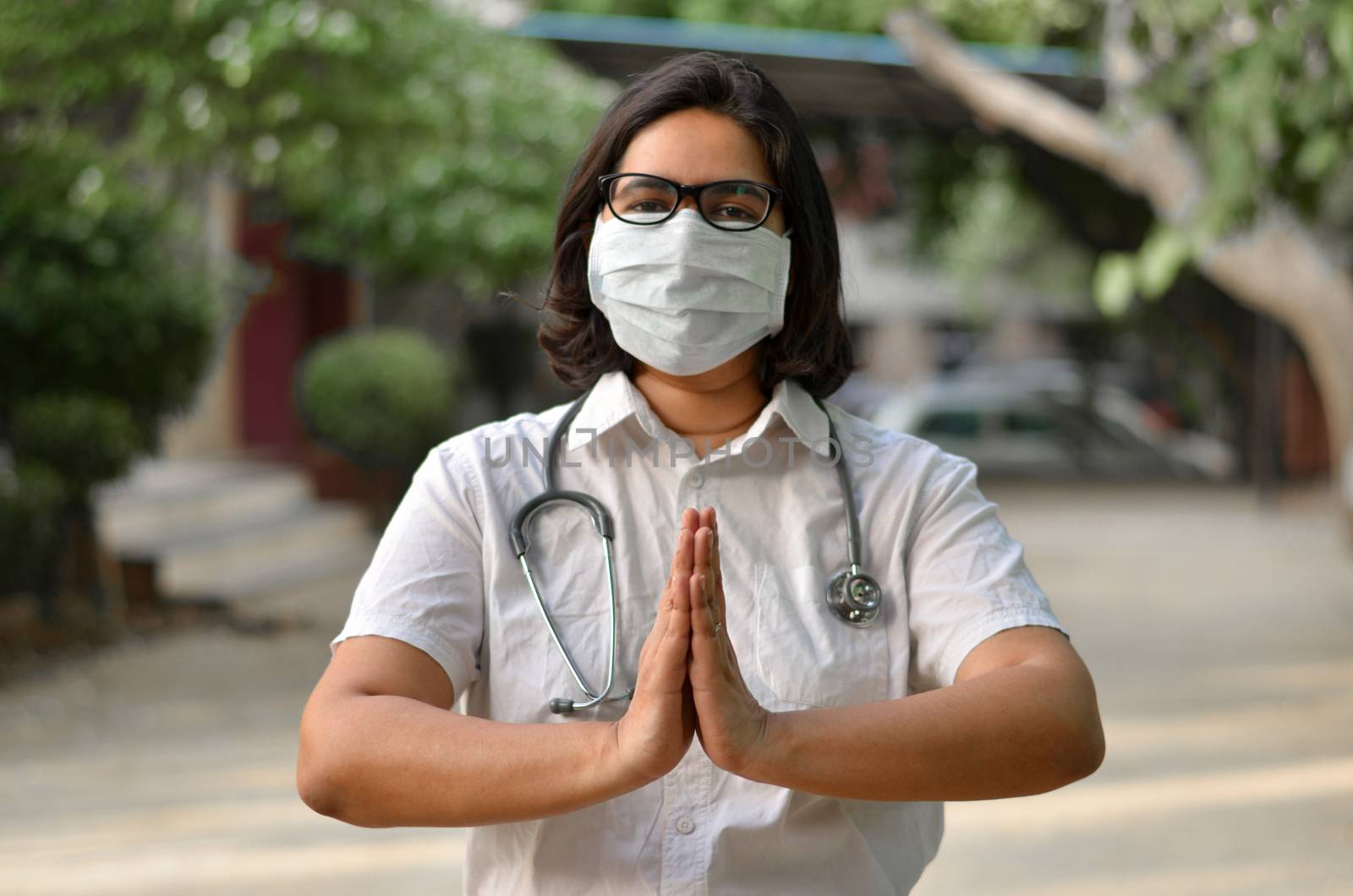 Portrait of a young medical healthcare female worker with hands crossed / folded in namaste, wearing surgical mask to protect herself from Corona Virus (COVID-19) pandemic against blur background
