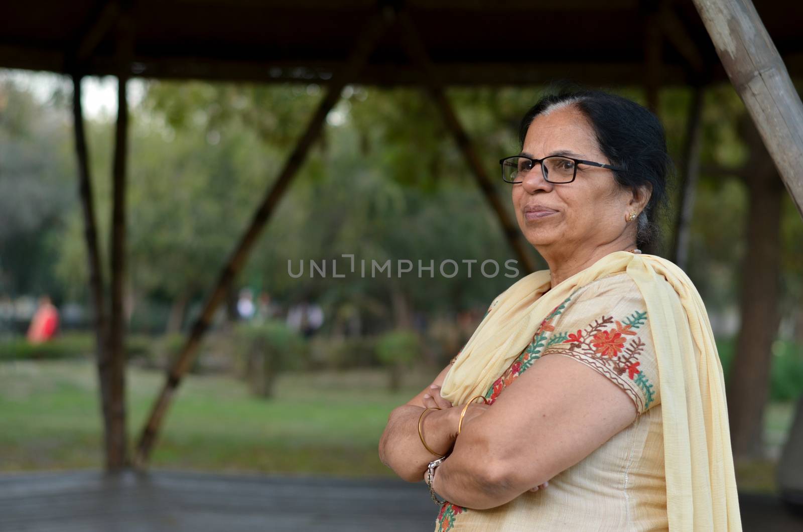 Smart and confident senior north Indian woman standing, thinking and looking away with hands crossed / folded in a park wearing an off white salwar kameez punjabi suit in summers in New Delhi, India