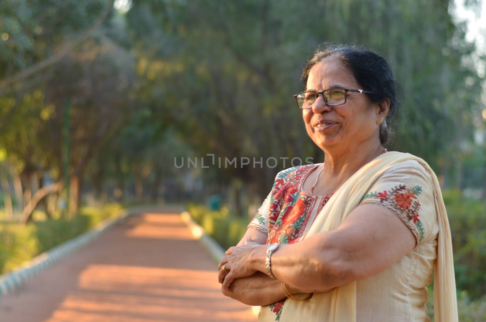 Smart and confident senior north Indian woman standing, thinking and looking away with hands crossed / folded in a park wearing an off white salwar kameez punjabi suit in summers in New Delhi, India