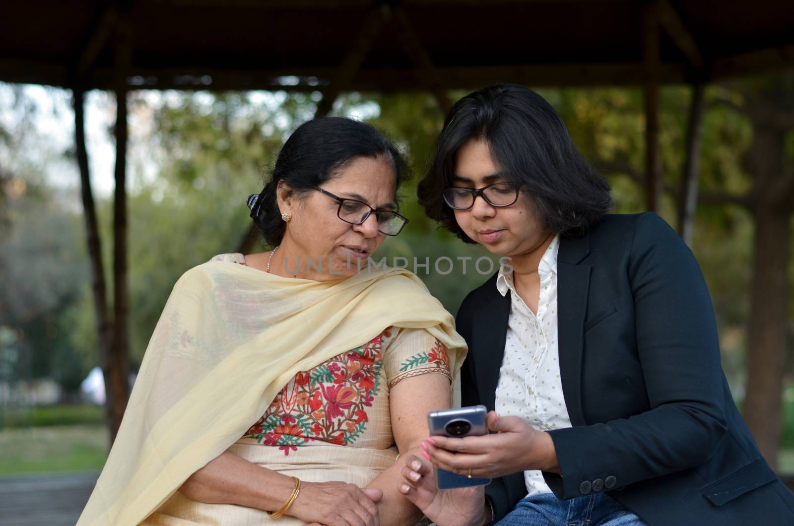 Smart young Indian girl with an old Indian woman looking at the mobile phone and busy talking sitting on a bench in a park in Delhi, India