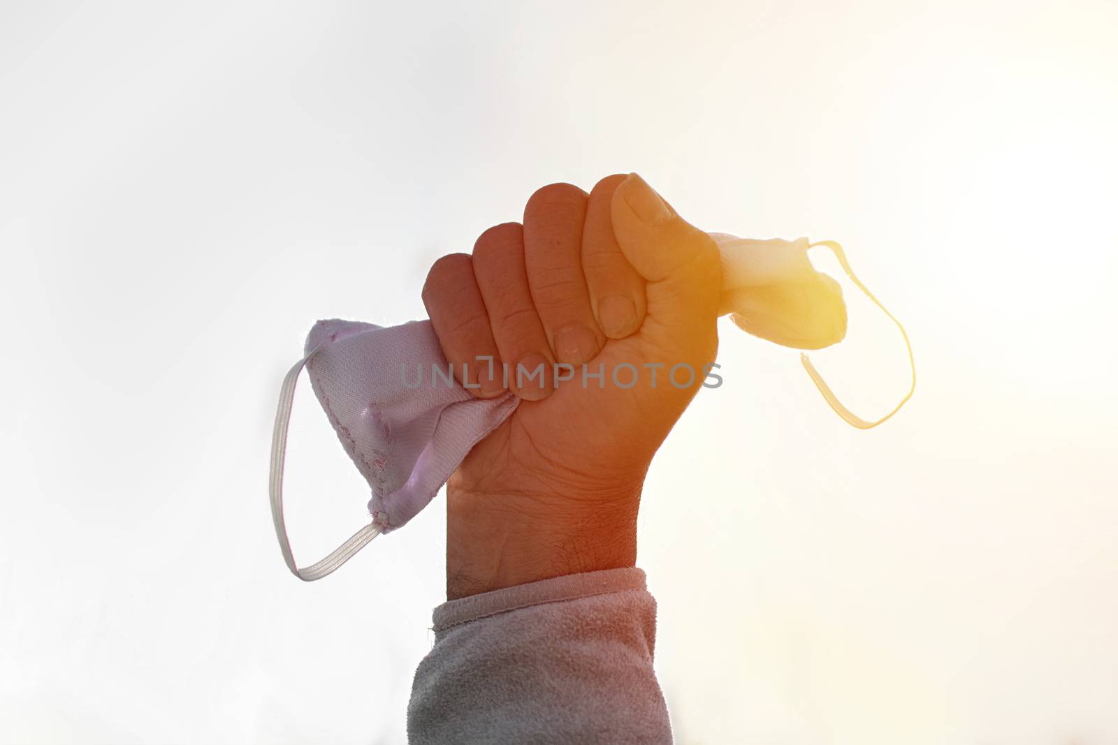 Victory over coronavirus concept. Cropped close up photo of male holding facial medical surgical blue mask in clenched fist arm isolated over white background at sunset with copy space.