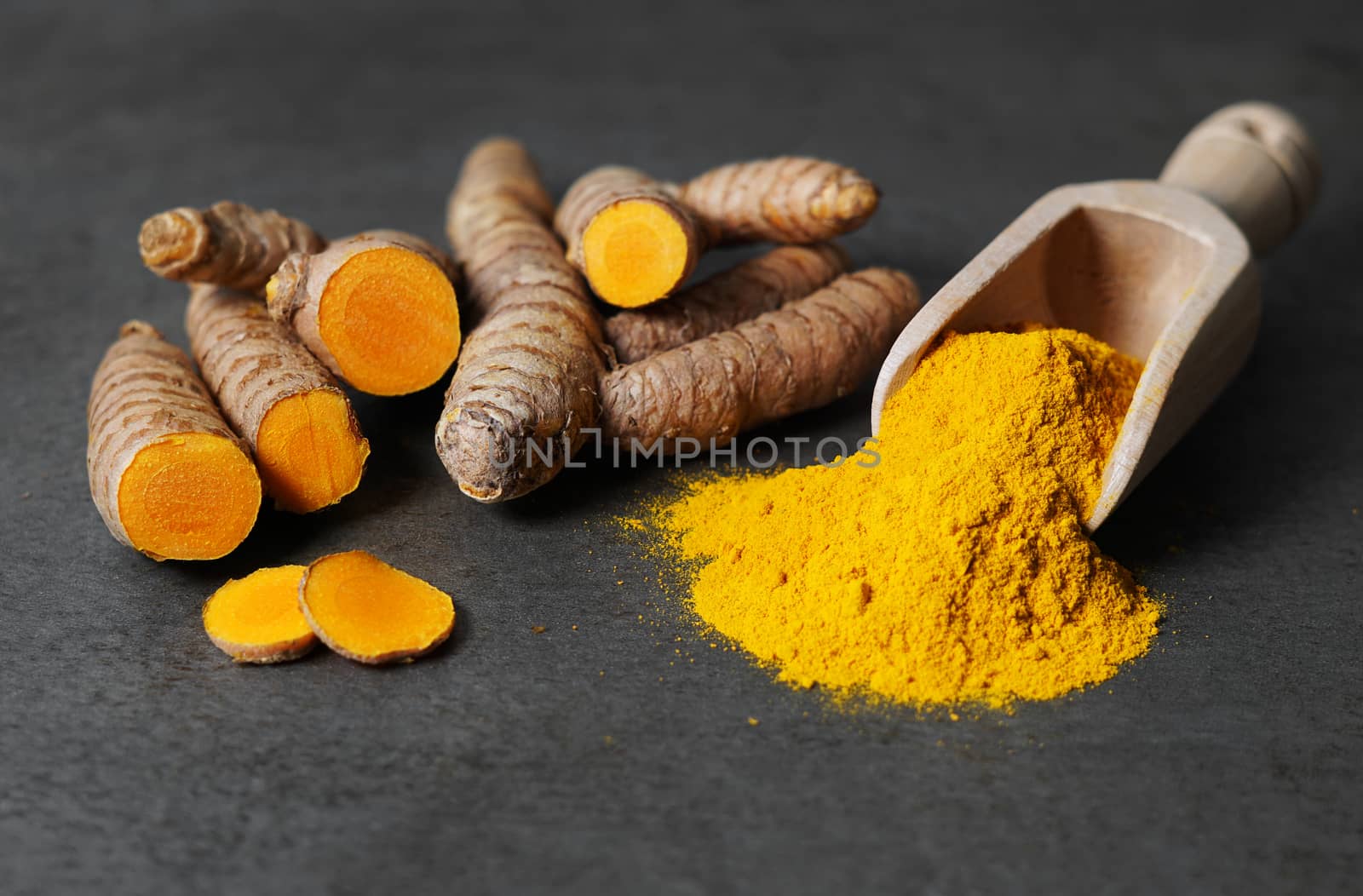 Turmeric powder healthy spice Asian food closeup of turmeric roots sliced and a wooden bailer on a rustic dark grey kitchen board.