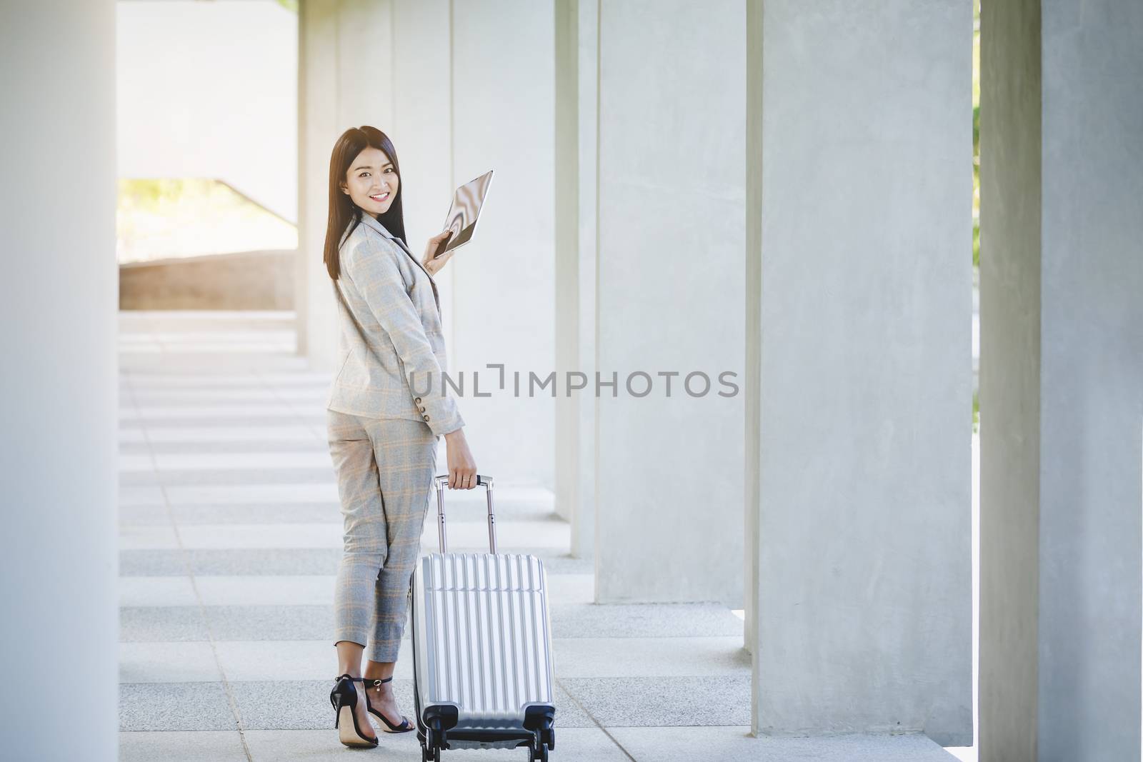 Portrait of business woman looking digital tablet with white travel bag on walkway while waiting to travel to the destination 