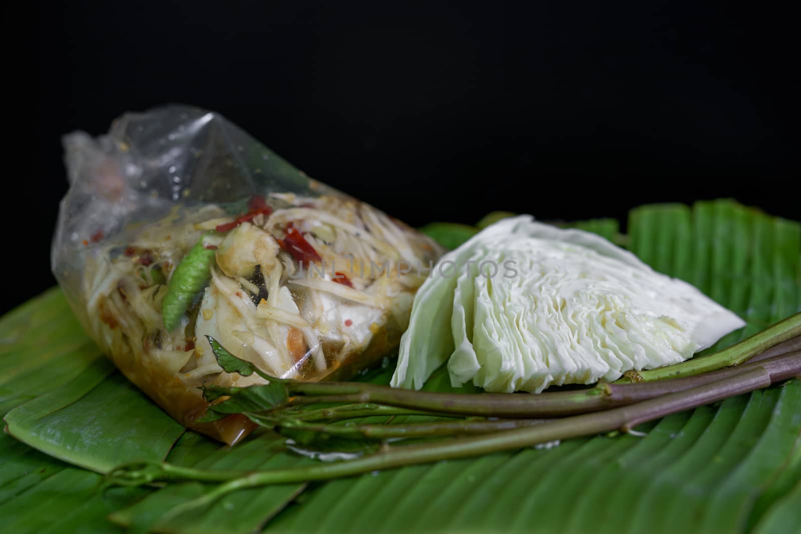 Thai food called papaya salad, pickled fish, salted eggs in a plastic bag and vegetables on a banana leaf, green, black background