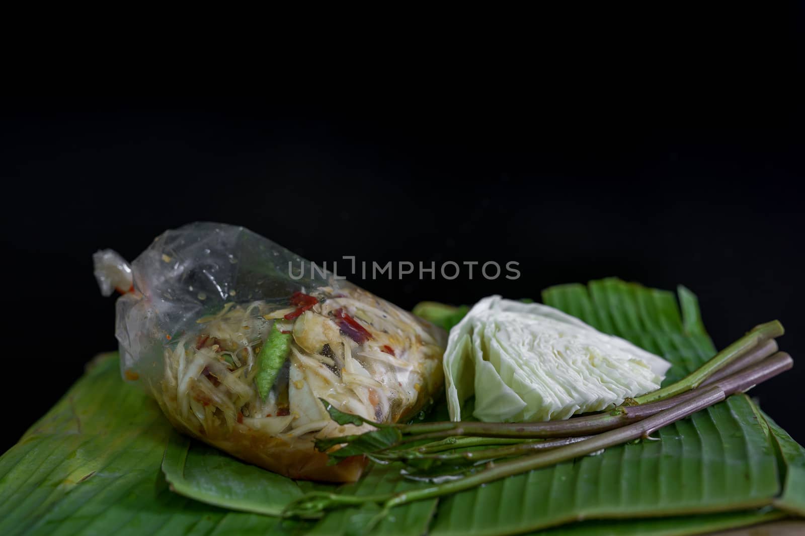 Thai food called papaya salad, pickled fish, salted eggs in a plastic bag and vegetables on a banana leaf, green, black background