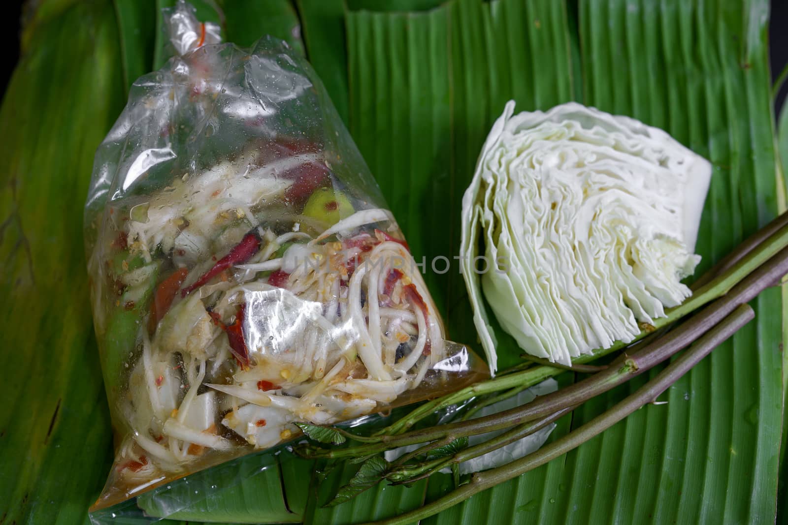 Thai food called papaya salad, pickled fish, salted eggs in a plastic bag and vegetables on a banana leaf, green, black background