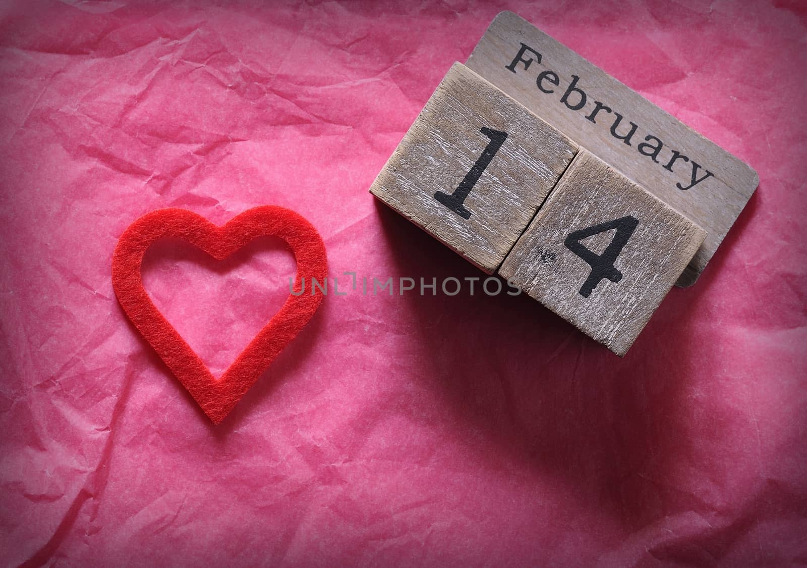 Wooden calendar with February 14th and red cut heart on pink paper. From above.