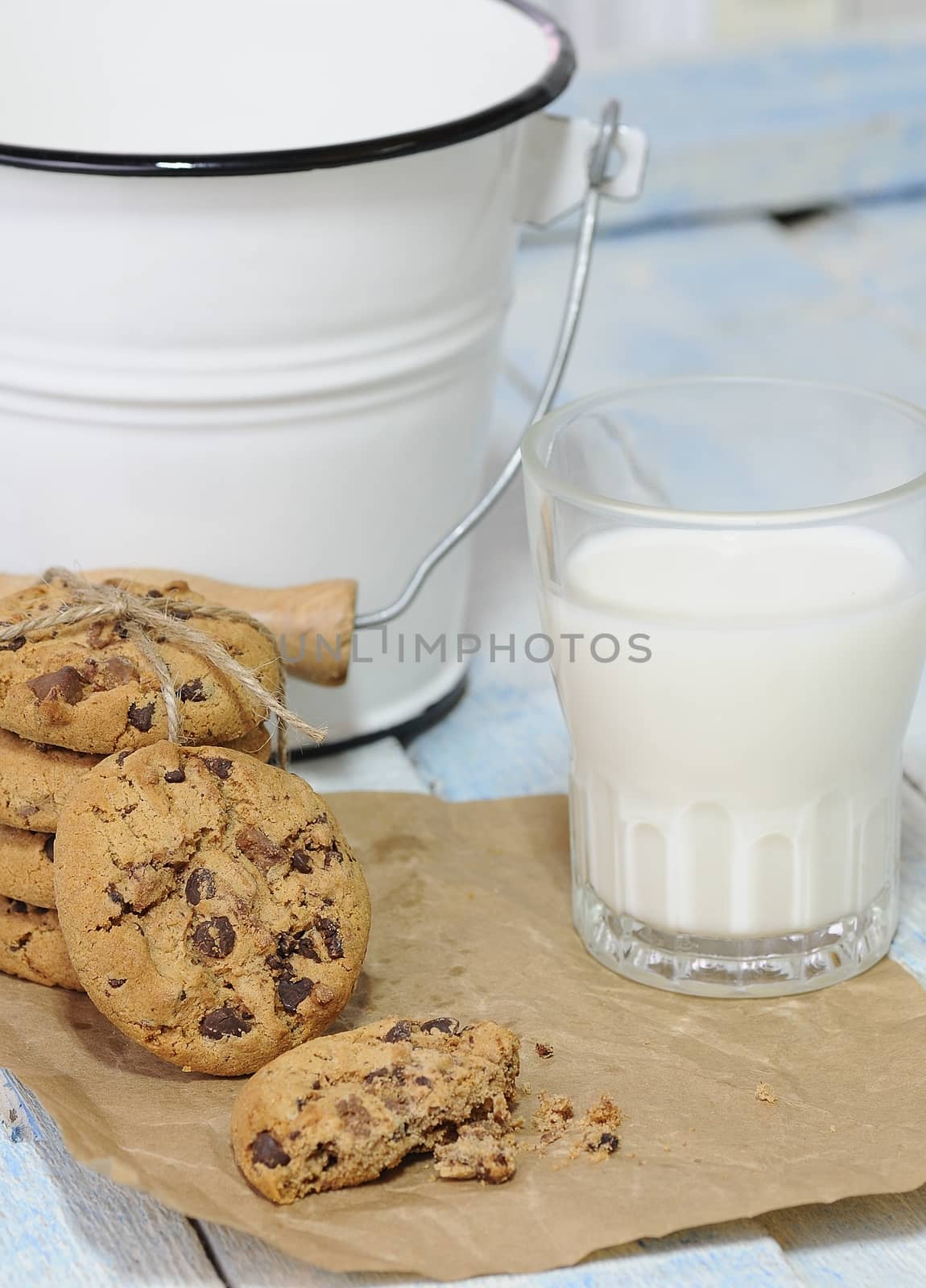 One bitten and stack of chocolate chip cookies with glass of milk on baking paper. Bucket on background.