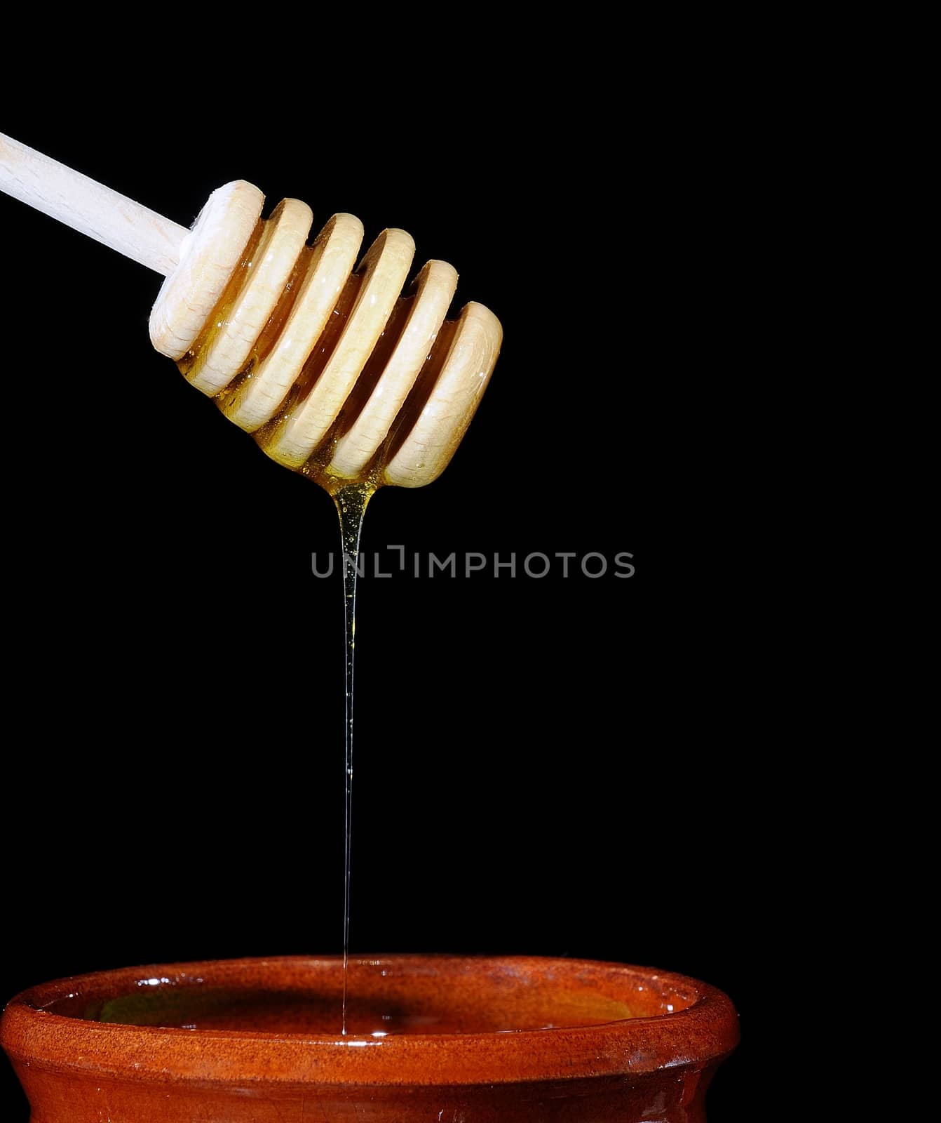 Close-up of wooden stick with liquid honey pouring in ceramic pot.Black background