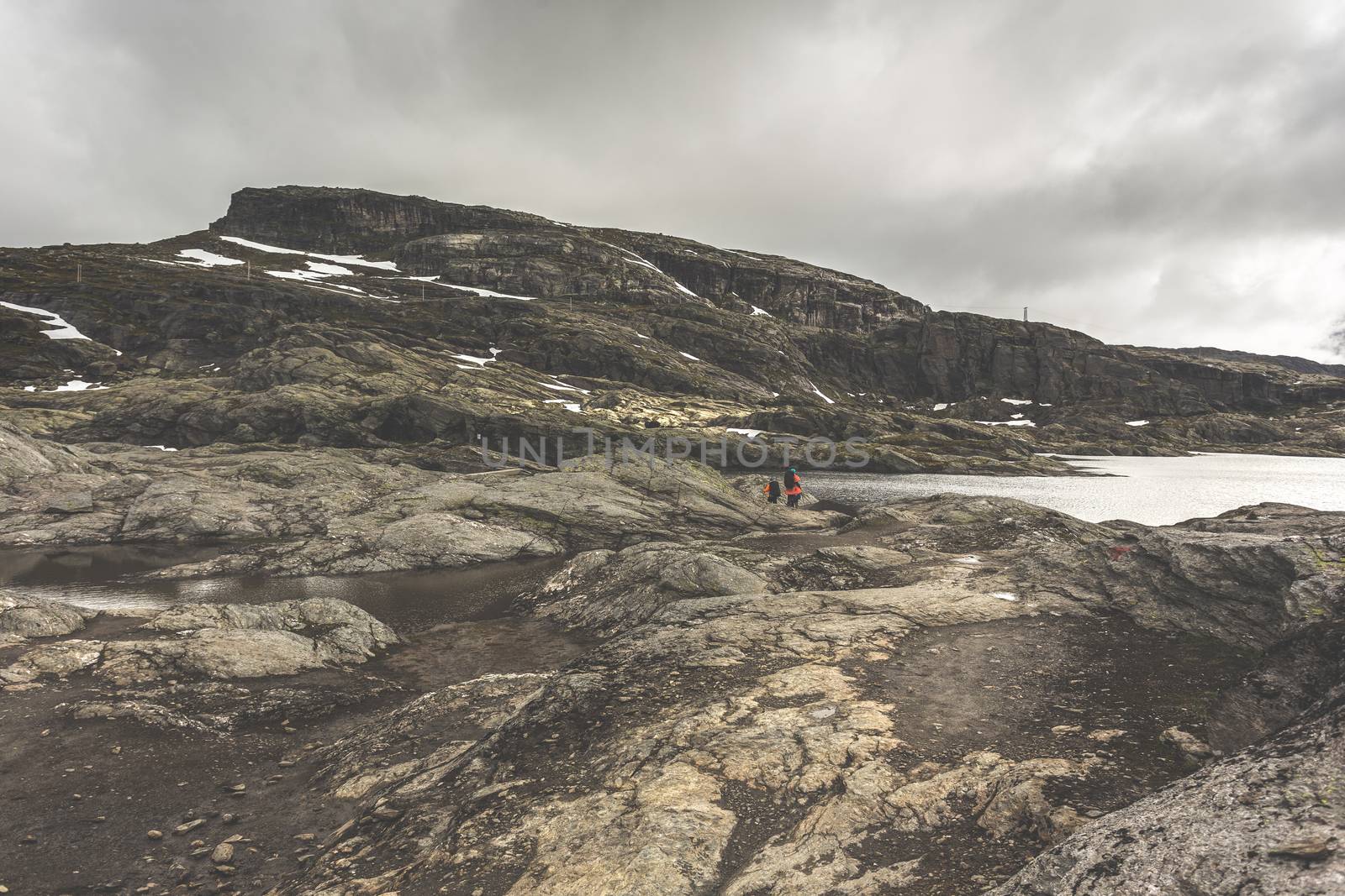 Tourists with backpacks walk across a rocky plain to the famous landmark - Troll tongue rock or Trolltunga. Cold and windy summer in Norway.
