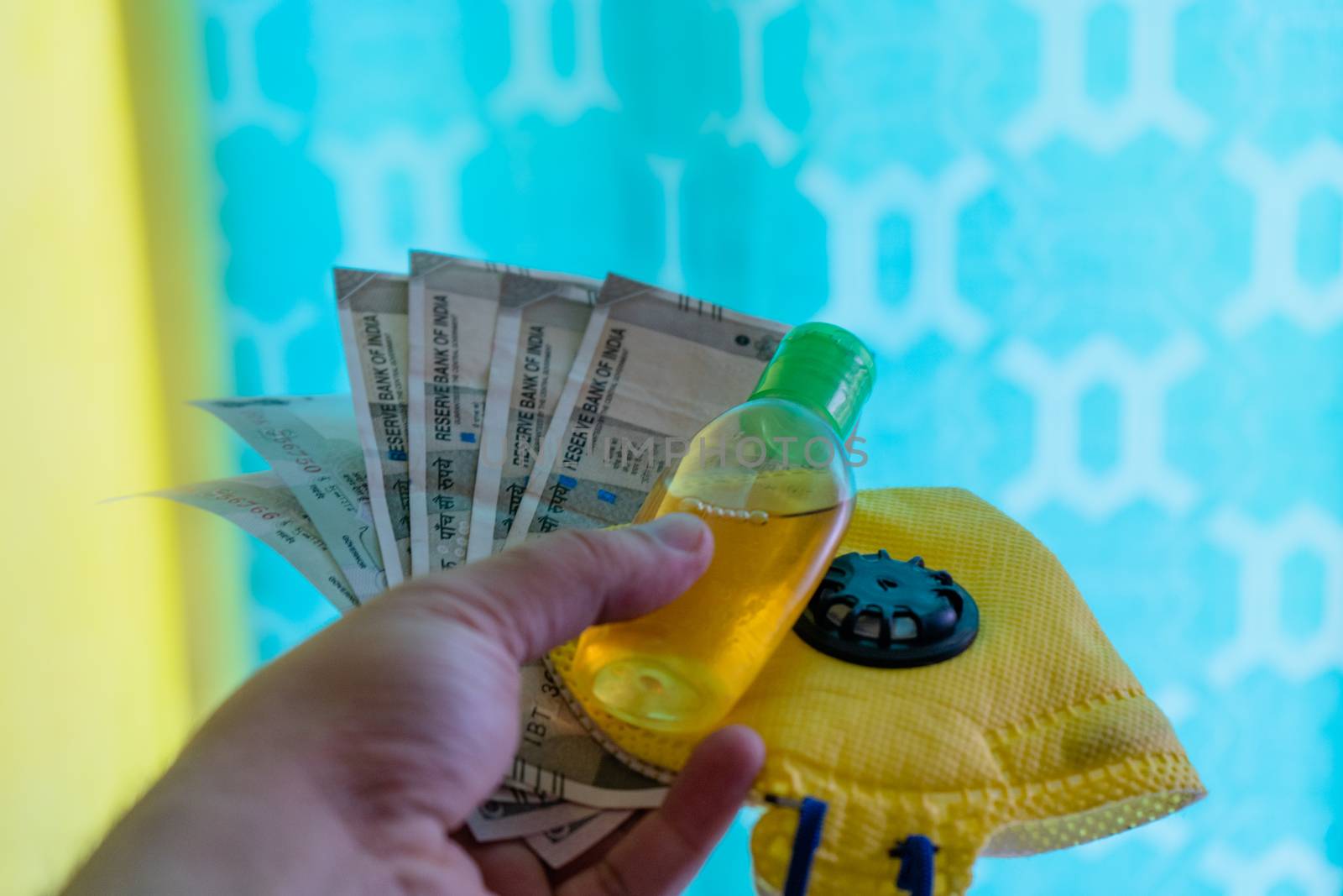 Hand holding out indian currency notes,yellow mask and sanitizer, against a colorful background by Shalinimathur