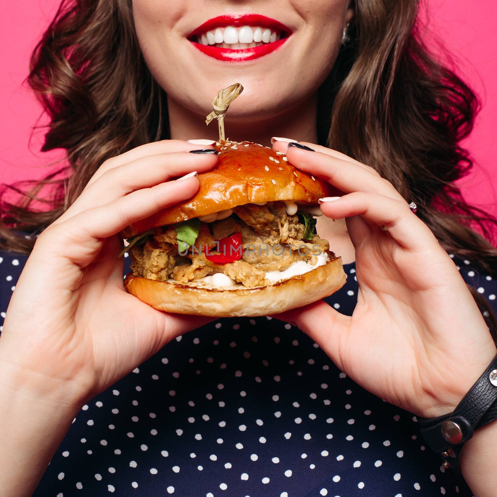 Close-up of smiling happy brunette woman with red lips holding delicious juicy burger with chicken, salad and vegetables in front of camera.