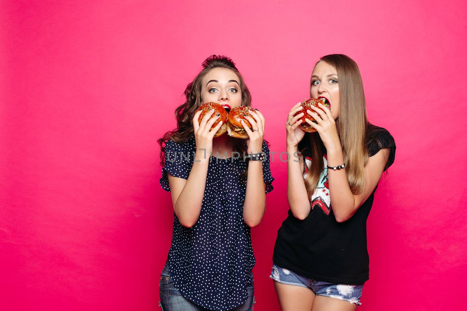 Studio portrait of girlfriends eating delicious and yummy burgers with chicken with hungry look at camera against vivid magenta background.