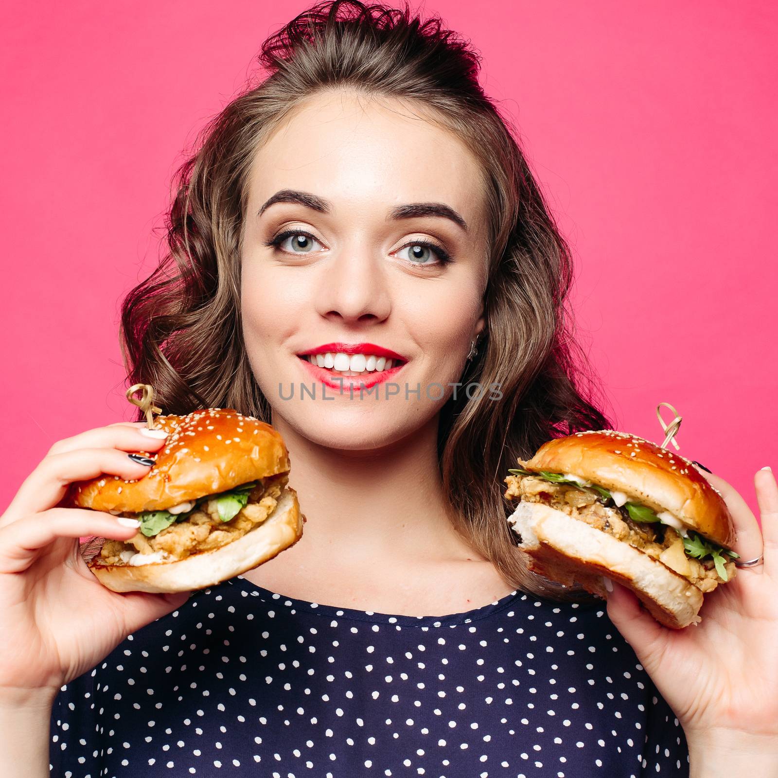 Beautiful positivity girl smilingat camera and posing with two tasty humburgers. Young pretty woman wearing in black holding big cheeseburgers. Pink studio background. Concept of fast food and diet.