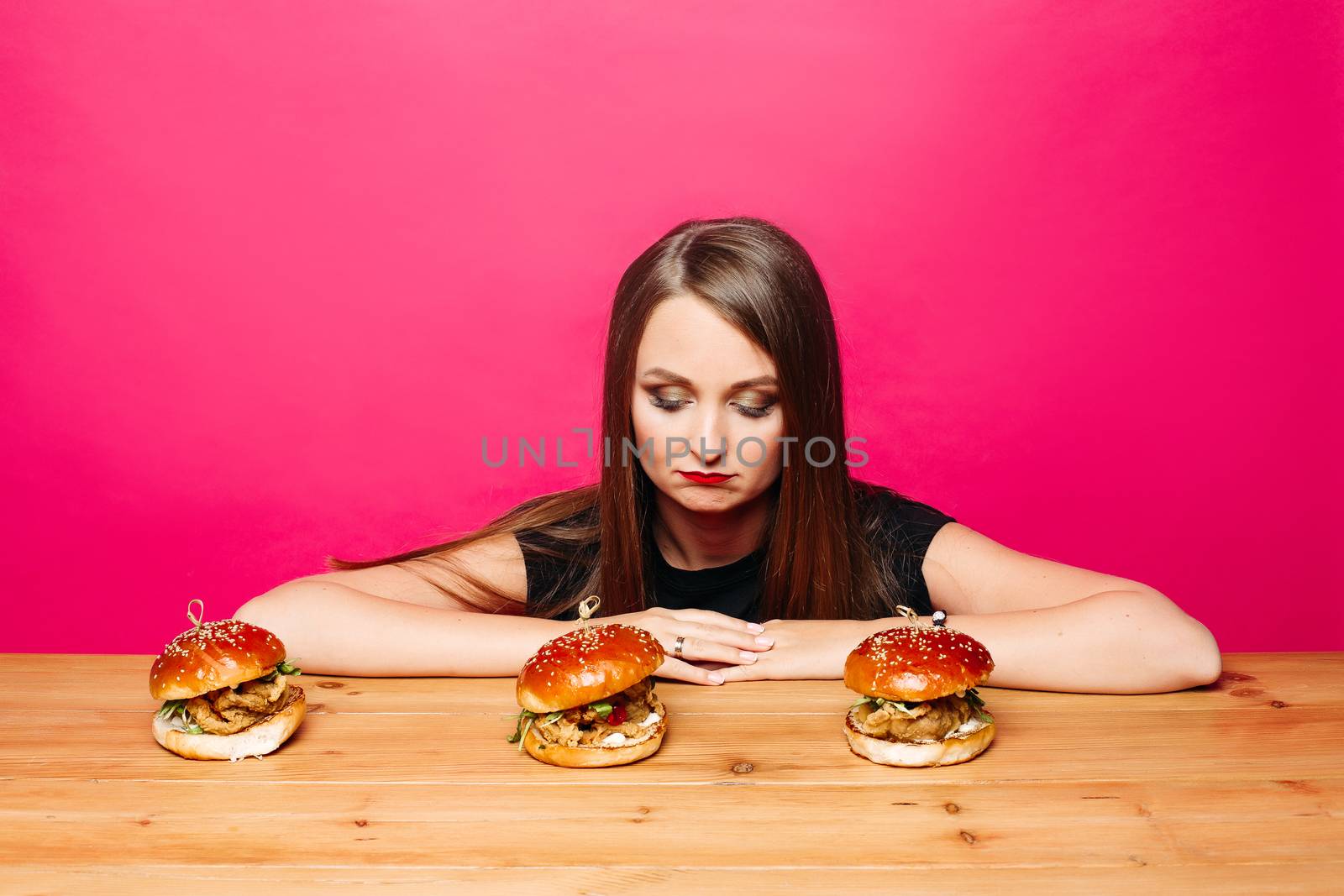 Portrait of unhappy pretty girl with hairstyle and make up looking at delicious freshly cooked hamburgers on wooden table with her chin on hands over bright magenta background.