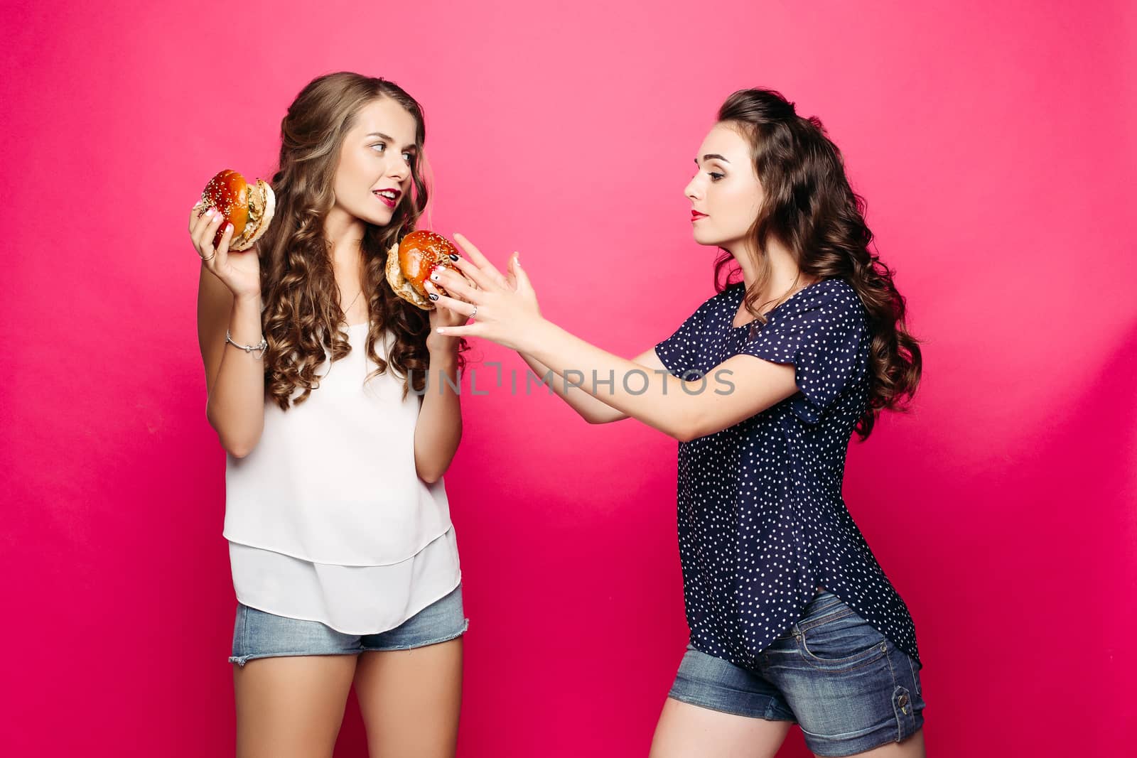 Give me my burger game between friends. Brunette girl pulling her arms towards her burger in friend s hand over pink background. Isolate.