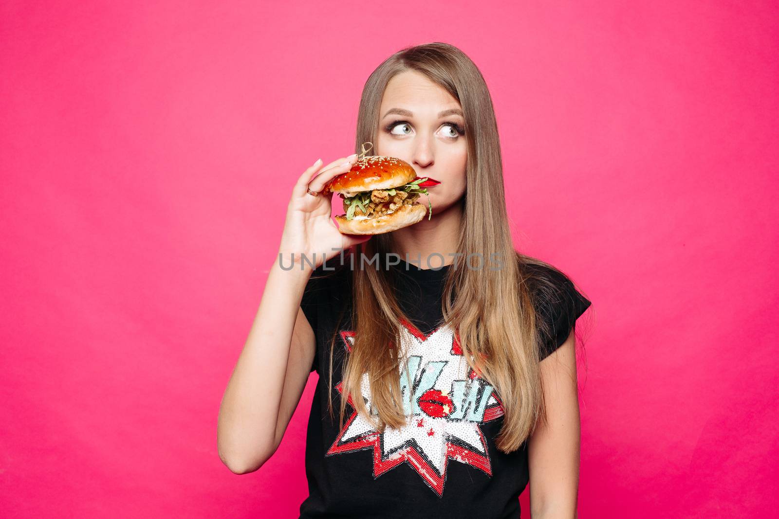Front view of sadly young girl looking at side and thinking eating tasty humburger or no. Pretty woman wearing in black t-shirt holding big cheeseburger, posing on pink background. Concept of diet.