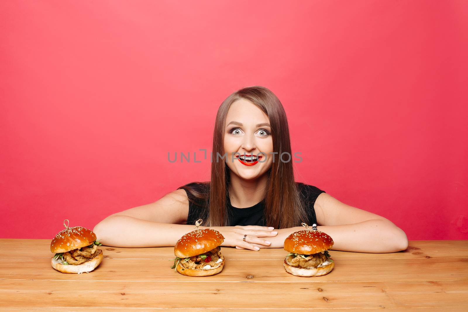 Studio portrait of excited woman with long straight hair looking at camera with open wide eyes sitting at table with three delicious burgers with chicken in front of her against red background.