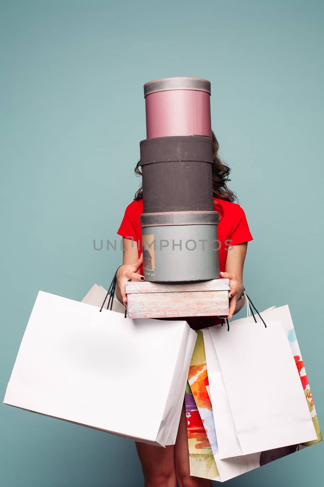 Studio portrait of incognito brunette woman in red dress holding at hands many bags, boxes and presents after shopping. Unrecognizable girl hiding against boxes. Sale in fashionable store.