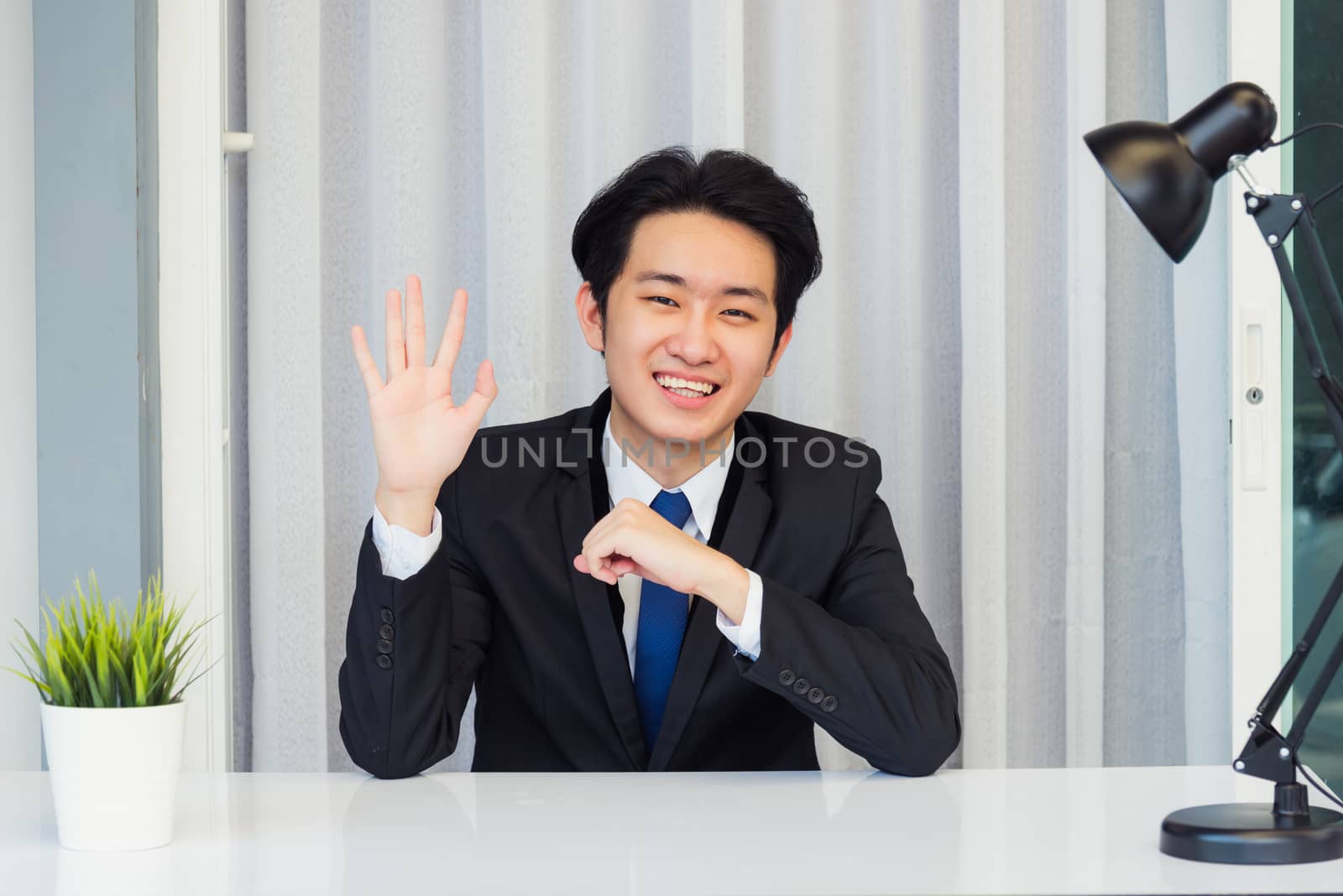 Work from home, Close up face of Asian young businessman video conference call or facetime he looking to camera sitting and raise your hand and say hello team or customer on a desk at the home office