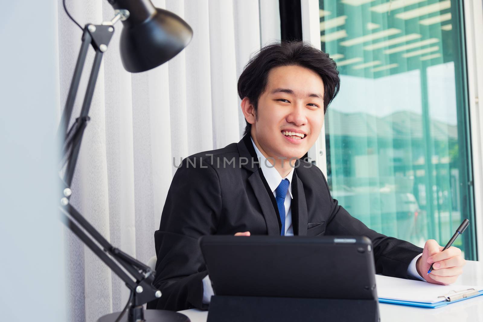 Work from home, Asian young businessman video conference call or facetime he smiling looking to camera sitting on desk using smart digital tablet computer and write notebook information at home office