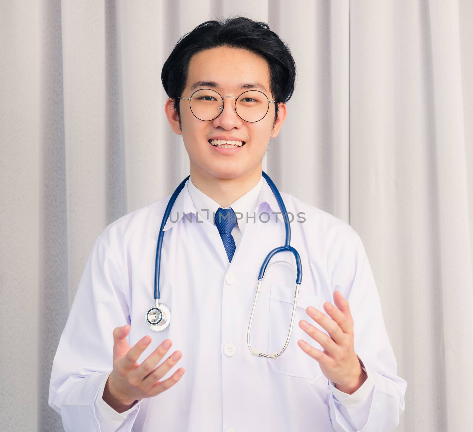 Portrait of Happy Asian young doctor handsome man smiling in uniform with stethoscope talking online video conference call or facetime raise his hand to explain, healthcare medicine concept