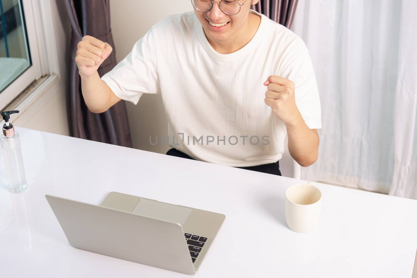 Happy Asian young business handsome man smile work from home office wear glasses, t-shirt comfortable exciting good news successful winner raises hand yes gesture with laptop computer on desk at home