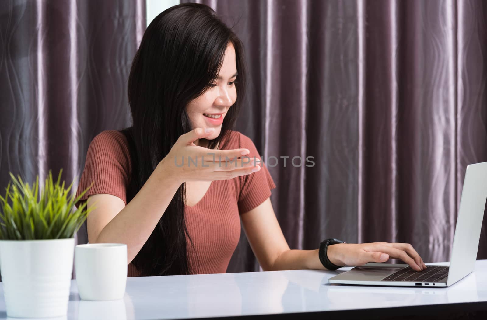 Work from home, Smiling Happy Asian business young beautiful woman sit on desk workspace video call conferencing by webcam laptop computer technology and raise your hand explain the job at home office