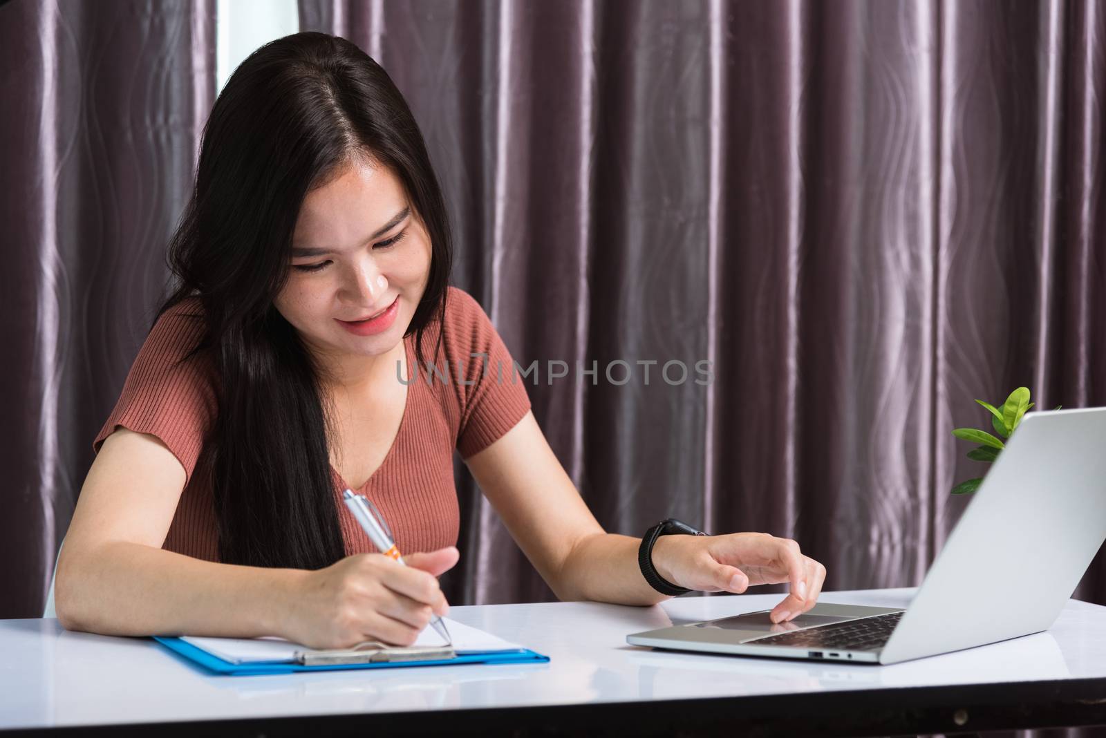 Work from home, Asian young businesswoman smile video conference call or facetime by laptop computer meeting with team and her writing on paper noted on desk at home office