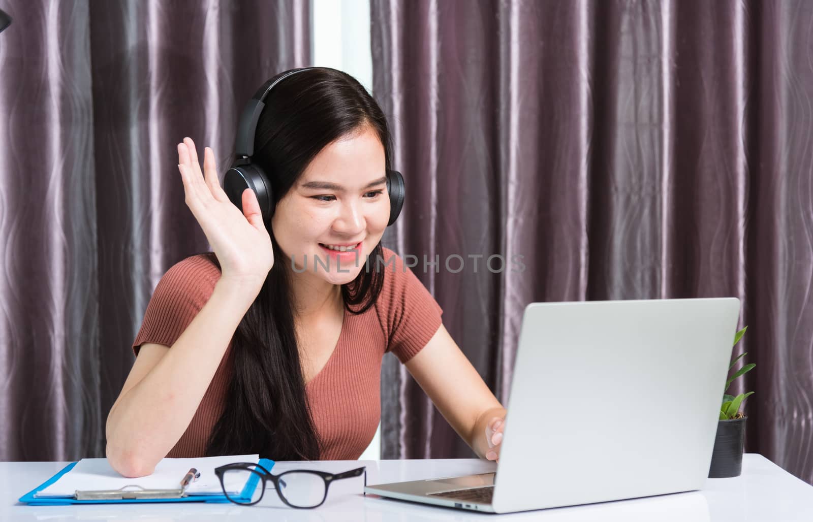 Work from home, Smiling Happy Asian business young beautiful woman sitting on desk workspace wearing headphones video call conference with laptop computer raise hand saying hi team at home office