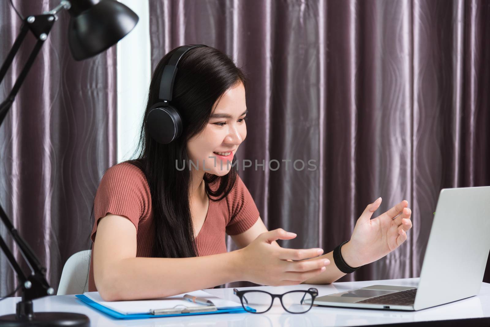 Work from home, Asian young businesswoman smile wearing headphones video conference call or facetime by laptop computer her raise his hand to explain to teammates on desk at home office
